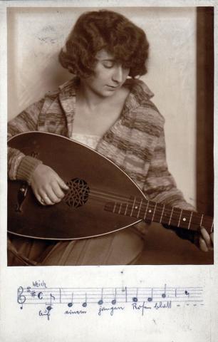 Black and white photograph of a young woman in three-quarter profile with wavy hair, which she wears as a bob. Her gaze is directed downward at the lute she is playing, so that her eyes appear closed.