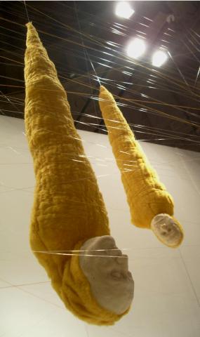 Two human figures in yellow cocoons hang upside down in a room from the ceiling, to which they are attached with transparent threads.