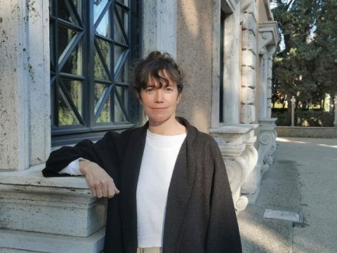 Dark-haired woman with white T-shirt and black jacket looks friendly into the camera. Her right arm leans on a window sill of an old stately building. 
