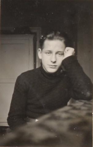 Schwarz-Weiß-Porträtfoto eines jungen Mannes, der direkt in die Kamera schaut. Er stützt den Ellbogen auf den Tisch auf, an dem er sitzt, seine Schläfe ist auf seiner linken, zur Faust geballten Hand aufgestützt