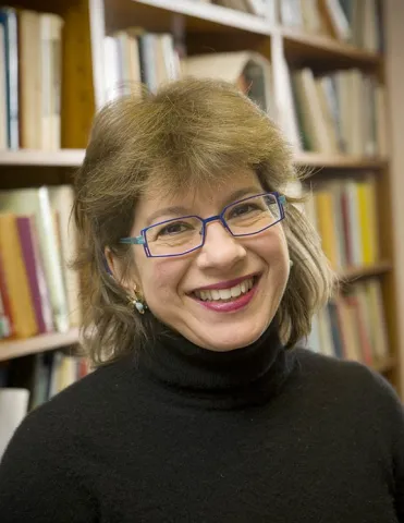 A blonde woman with blue glasses and a black turtleneck sweater stands in front of a bookshelf and smiles into the camera.