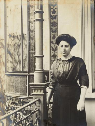 Schwarz-Weiß-Porträtfoto einer jungen Frau. Sie trägt ein hochgeschlossenes Kleid im Stil der 1910er Jahre und eine Hochsteckfrisur und steht vor einem Haus, dessen prächtige Fassadenverzierung im Hintergrund zu sehen ist.