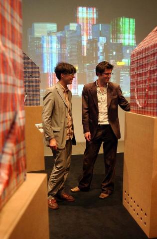 Two men between houses made of checkered plastic bags on cardboard boxes, in the background a projection