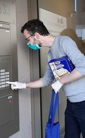 A man wearing a surgical face mask with a package of matzo under his arm rings a doorbell.