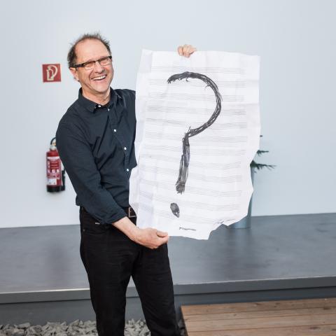 Paul Brody hält ein großes Porträt von Notenblättern mit einem Fragezeichen darauf.
