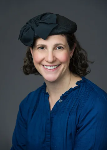Porträt einer in die Kamera lächelnden jungen Frau mit schulterlangen dunklen Locken, schwarzem Hut und blauer Bluse.
