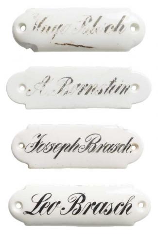 Three white porcelain signs one below the other with the names Hugo Bloch, A. Bernstein, Joseph Brasch and Leo Brasch written in cursive.