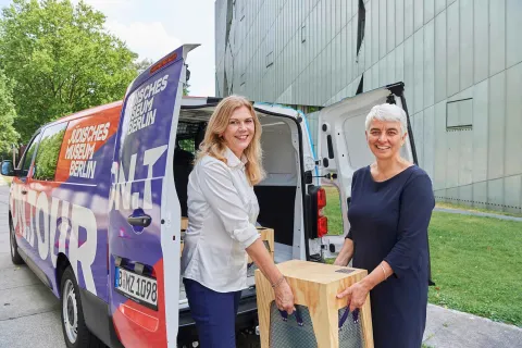 Eine blonde Frau in wei&szlig;er Bluse und eine grauhaarige Frau in dunkelblauen Kleid halten eine Kiste aus Holz vor den aufgeklappten T&uuml;ren eines Transporters.