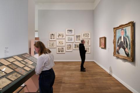 Exhibition space with visitors and paintings on white wall, a showcase with paintings and drawings in the center of the room.