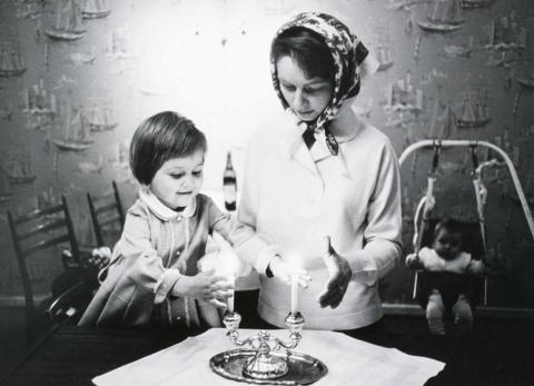 Ein kleines Mädchen (links) und eine junge Frau mit Kopftuch (rechts) halten ihre Hände neben die Flammen zweier Kerzen, die in einem zweiarmigen Kerzenständer auf einem Tisch mit weißer Tischdecke stehen