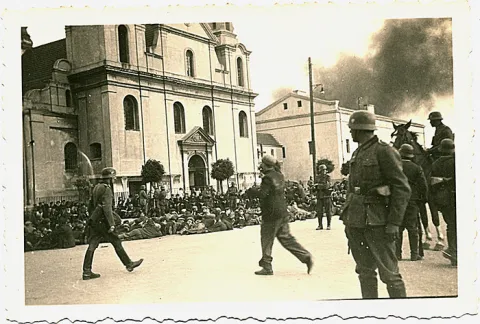 Schwarz-wei&szlig; Fotografie von einer Kirchenfassade vor der Menschen auf dem Boden sitzen, im Vordergrund deutsche Soldaten.