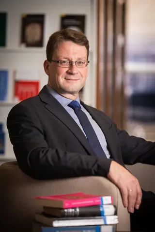 A man with dark hair, glasses, in a suit and tie looks friendly into the camera.
