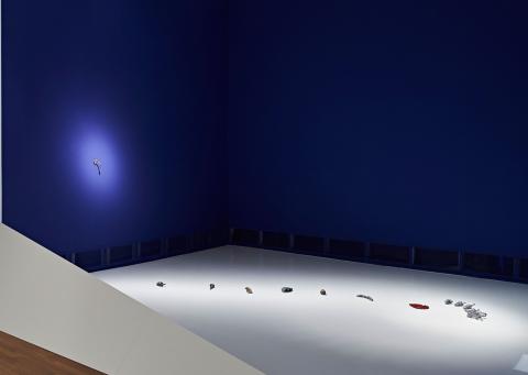 Some small objects lie on a white surface in a sparsely lit room with dark blue walls. The objects and a circular small area on one wall are specifically illuminated with spotlights.