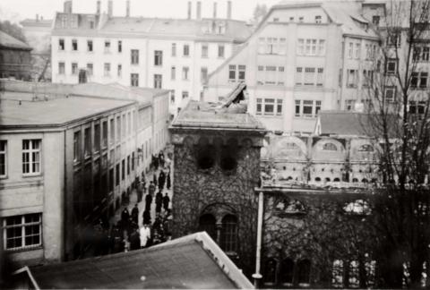 The photo shows a destroyed synagogue whose roof is missing, it seems to have been burnt, rubble lies on the still existing entrance portal, in the street in front of it there are many passers-by. 