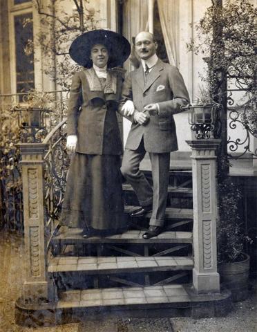 A woman and a man are standing together on a staircase in front of a house. She is wearing a huge hat and gloves with a dark dress and has hooked up with him, he is wearing a suit with a pocket square.
