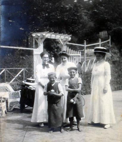 Drei Damen in weißen Kleidern, eine mit großem Hut, stehen vor einem Eingangstor. Vor ihnen stehen zwei barfüßige Jungs mit Tennisbällen in den Händen.