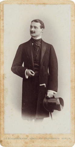 Black and white portrait photo of a standing young man. The body is seen frontally, the head turned to the left in three-quarter profile. He wears a mustache and a frock coat, in his hand he holds a top hat and a walking stick.