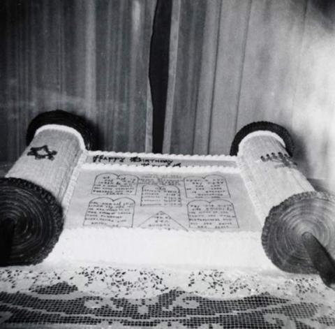 Black-and-white photo of a cake shaped like a Torah scroll on a table with a lace tablecloth