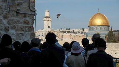 A group of people are standing in the Old City of Jerusalem looking at a dove in the sky. On the right side of the picture you can see the golden dome of the Dome of the Rock.