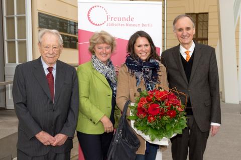 Photo, left to right: W. Michael Blumenthal, Monika Grütters, Paula Konga, and Peter Schäfer smile at the camera and hold a large display of red flowers