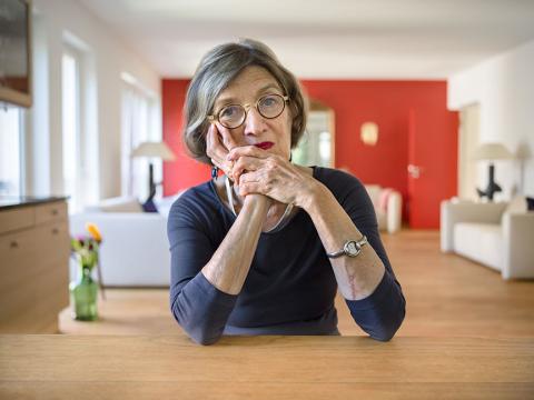 Eine Frau sitzt an einem Tisch, das Kinn auf die Hände gestützt, im Hintergrund ein weitläufiger Wohnraum