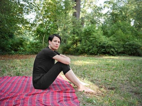 A woman dressed in black with short hair sits on a red picnic blanket on a meadow and looks into the camera
