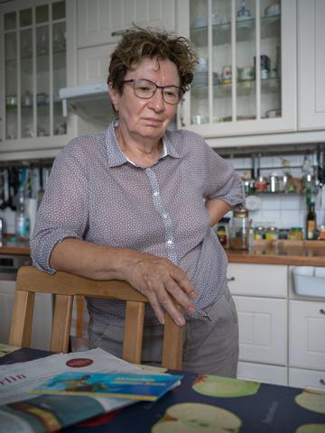 A woman with glasses and dotted blouse stands leaning on a chair back in a kitchen