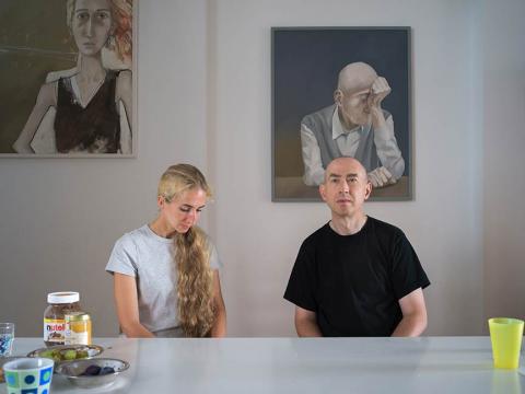 A woman with long blond hair and a bald man, both wearing T-shirts, sitting at a table, cups and a Nutella glass can be seen in the front left, two modern paintings in the background