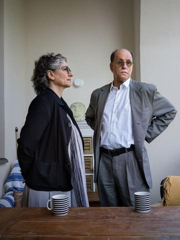 A couple dressed in shades of black, white and gray stand behind a table with striped tea cups, she in half-profile looking to the right, he frontally but not looking directly at the camera