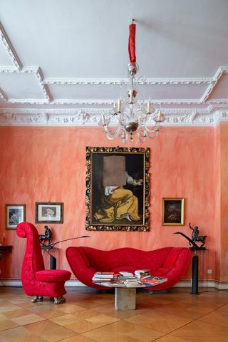 View of a room with stucco ceiling, chandelier, red designer couch set and above the sofa on the reddish painted wall a gold framed old oil painting of a woman with a rectangular piece of canvas missing at the level of the head