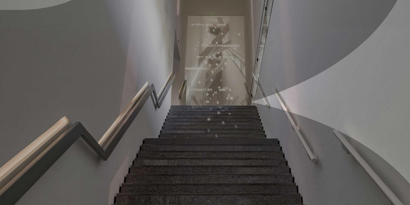 Treppe in einem Neubau, an deren Ende eine Projektion mit weißen Wörtern in Hebräisch und Deutsch. Das Bild ist mit einem grauen Schleier der das JMB-Logo andeutet.