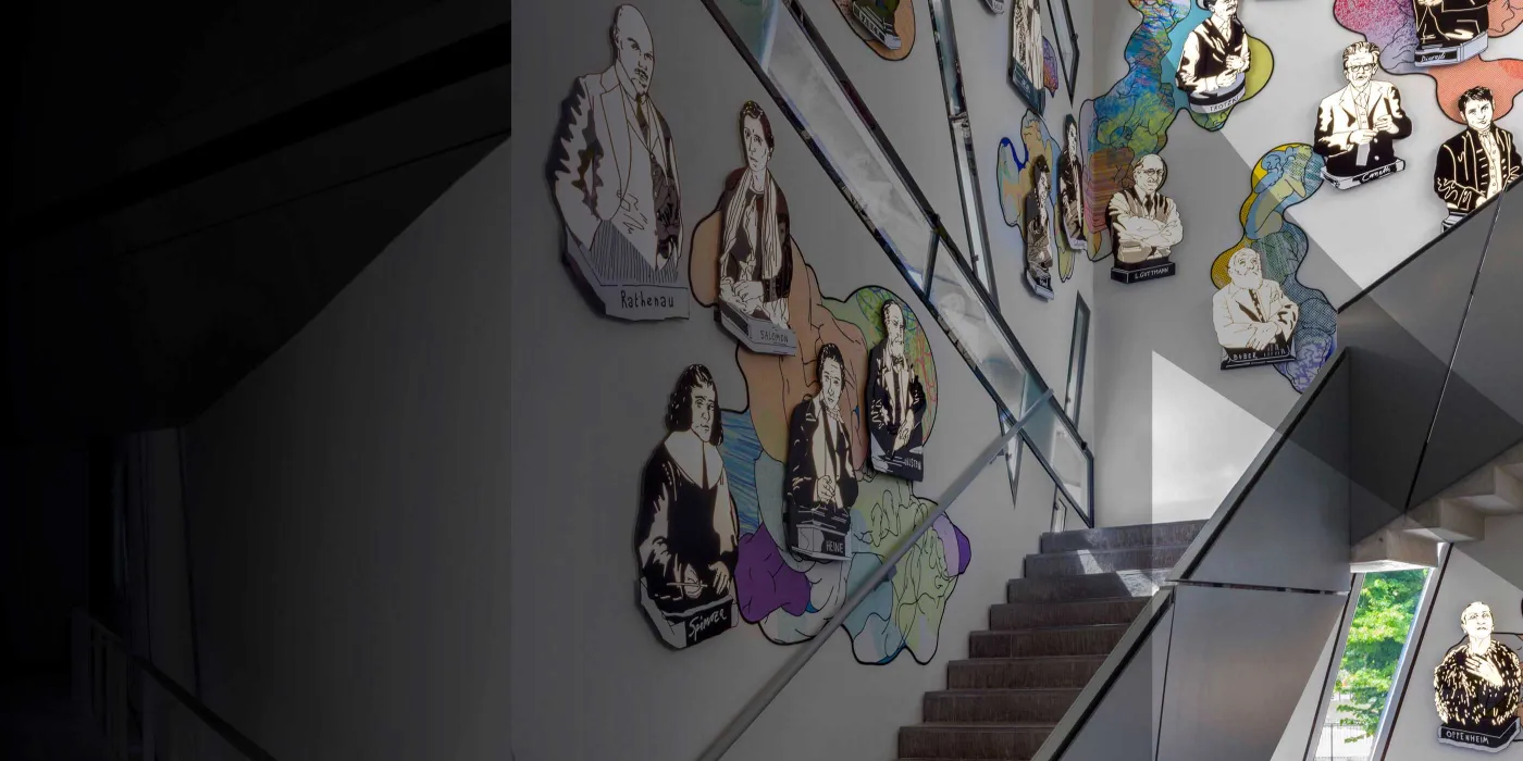 Schwarze Treppe. An der Wand bunte Porträts. Das Bild ist mit einem grauen Schleier der das JMB-Logo andeutet.