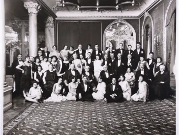 Black-and-white photo. Large group photo of a festive gathering. The ladies and gentlemen are wearing elegant clothing and are standing in a large room.