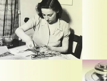 A woman is sitting at her desk covered with colours and brushes. She is painting.