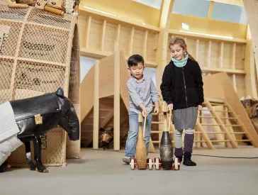Zwei Kinder schieben jeweils eine Spielente am Stab durch das Kindermuseum ANOHA. Ein Tapirskulptur links im Bild scheint ihnen zuzuschauen.