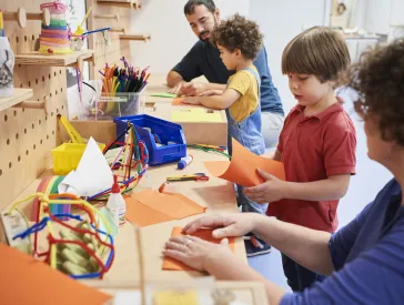 Zwei Kinder und zwei Erwachsenes basteln mit Papier an einer Werkbank.