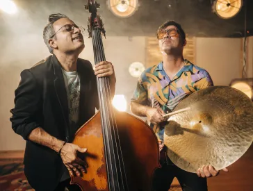 One man is playing bass, while the other is holding a cymbal in his hand. Both have their eyes closed. Spotlights are visible in the background.