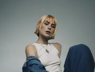 Frau mit kurzen Haaren und Pony sitzt auf dem Boden, tr&auml;gt Jeans und ein wei&szlig;es Tanktop und schaut in die Kamera.