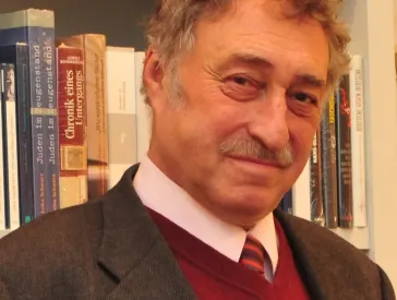 A middle-aged man stands in front of a bookshelf and looks into the camera.