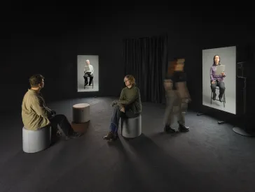 Zwei Menschen sitzen sich gegen&uuml;ber in einem dunklen Ausstellungsraum. Eine Person l&auml;uft durch den Raum. An den Wandseiten des Raumes sind zwei gro&szlig;e Monitore auf denen Menschen zu sehen sind, die auf St&uuml;hlen sitzen. 