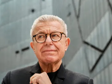 Portait Foto von Daniel Libeskind. Im Hintergrund ist der Libeskind Bau des J&uuml;dischen Museum Berlin unscharf zu sehen.