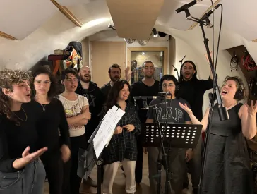 Eine Gruppe von Leuten steht zusammen in einem Raum. Sie singen zusammen. Vor ihnen stehen Notenst&auml;nder und Mikrofone. 