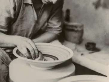  Black and white photograph of a woman working on a pottery bowl at a potter's wheel.