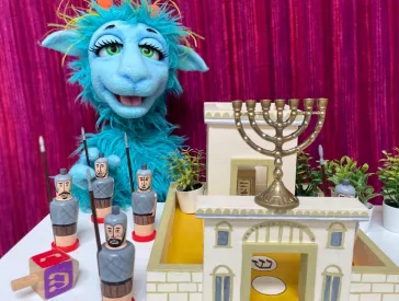 Eine blaue zottelige sprechende Puppe mit großen schönen Augen hinter einem Tisch mit Spielzeugfestung, Dreidel und Chanukka-Leuchter darauf.