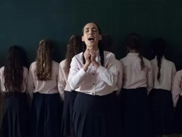 Mehrere junge Frauen stehen in einer Reihe mit dem R&uuml;cken zur betrachtenden Person. Eine junge Frau steht vorne, leicht vor der Reihe, frontal zur Kamera. Sie hat die Augen geschlossen, den Mund ge&ouml;ffnet und h&auml;lt ihre H&auml;nde senkrecht vor sich, leicht zu F&auml;usten geballt, sodass sie sich ber&uuml;hren.