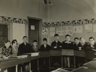 Das Bild zeigt einen Schulklassenraum mit elf Sch&uuml;lerinnen und Sch&uuml;lern, die in einem Halbkreis angeordnet an ihren Pulten sitzen.