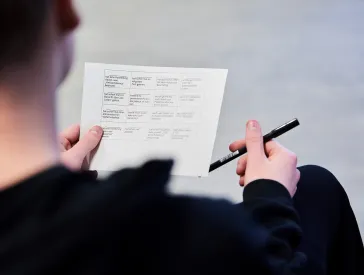 View over the shoulder of a person holding a worksheet in their left hand and a pen in the other.