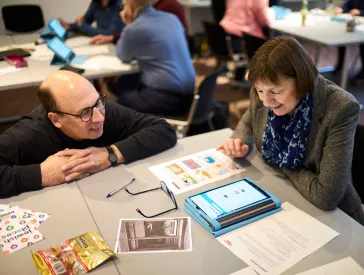 Zwei Personen sitzen an einem Tisch in einem Workshop- oder Seminarraum und betrachten gemeinsam Unterlagen sowie ein Tablet. Auf dem Tisch liegen ausgedruckte Materialien, eine Brille und weitere Arbeitsunterlagen. Im Hintergrund arbeiten weitere Teilnehmende an Tischen.