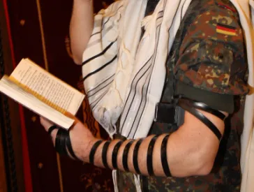 Ein Mann in Bundeswehruniform h&auml;lt in der linken Hand ein aufgeschlagenes Buch, die rechte Hand h&auml;lt er vor seine Augen, sein Blick ist gesenkt auf das Buch. An seinem linken Arm tr&auml;gt er einen j&uuml;dischen Gebetsriemen.
