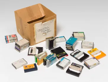 Ein leerer, alter Karton steht auf weißem Untergrund. Um den Karton liegen etwa 30 Audiokassetten in handbeschrifteten Hüllen. 